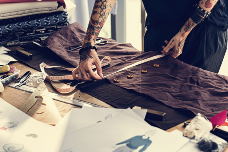 This is an image showing a person measuring a garment on a design table that has drawing tools, drawings, the garment being measured and some textile samples