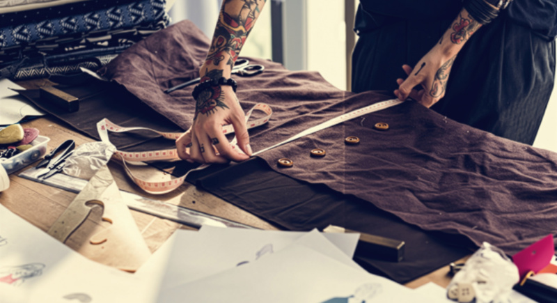 This is an image showing a person measuring a garment on a design table that has drawing tools, drawings, the garment being measured and some textile samples