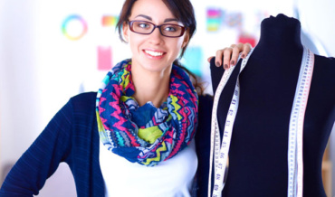 Fashion designer posing with a black dress form with many tape measures hanging around the dress form's neck